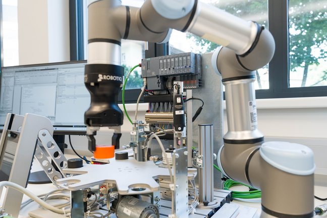 A collaborative robot arm with a gripper operates above a test setup on a lab bench; in the background are control units, cables, and a monitor in front of a window.