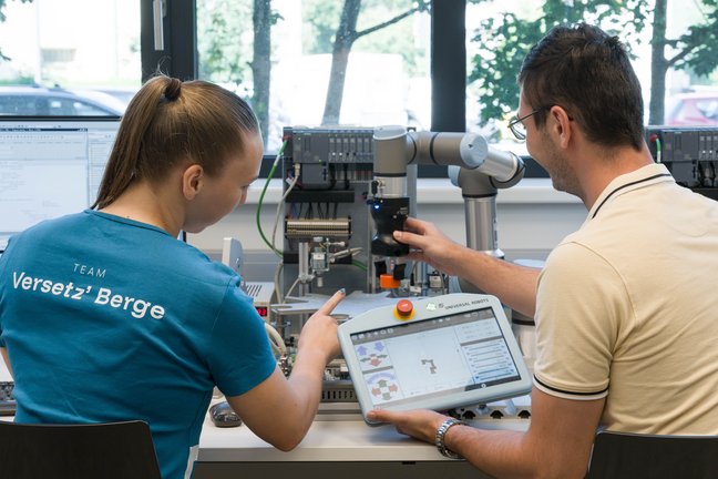 Two people sit at a lab bench operating a collaborative robot arm; one holds a teach pendant with a control interface while the other points at the test setup. In the background are control units, cables, and a window; a T-shirt reads “Team Versetz’ Berge”.