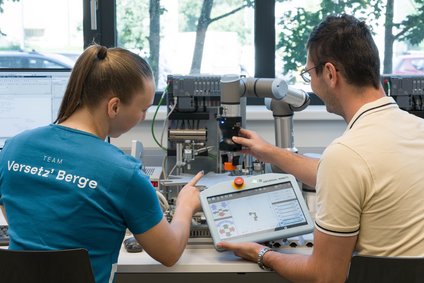 Zwei Personen sitzen an einem Labortisch und bedienen einen kollaborativen Roboterarm; eine Person hält ein Teach-Pendant mit Steueroberfläche, die andere zeigt auf den Versuchsaufbau. Im Hintergrund stehen Steuergeräte, Kabel und ein Fenster; auf einem T‑Shirt steht „Team Versetz’ Berge“.