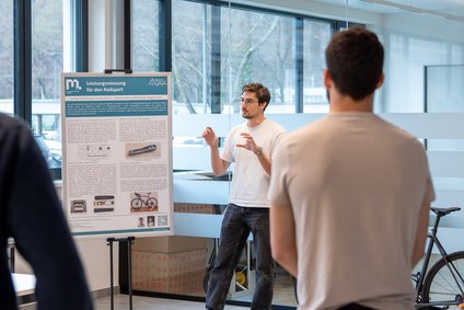 Person präsentiert ein Poster „Leistungsmessung für den Radsport“; Zuhörer im Vordergrund, Fahrrad rechts.