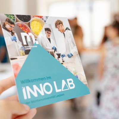 Hand hält einen Flyer „Willkommen im INNO LAB der Montanuniversität Leoben“; im Hintergrund unscharf eine Gruppe bei einer Veranstaltung.