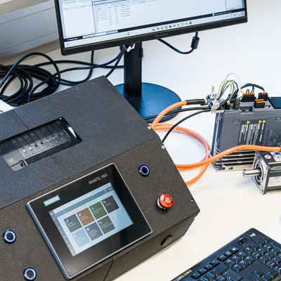 Laboraufbau zur Automatisierung: Monitor mit TIA‑Portal, HMI‑Bedieneinheit/PLC mit Touchscreen, Tastatur und verkabeltes Antriebssystem.