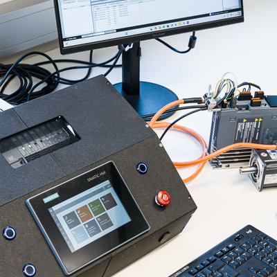 Laboraufbau zur Automatisierung: Monitor mit TIA‑Portal, HMI‑Bedieneinheit/PLC mit Touchscreen, Tastatur und verkabeltes Antriebssystem.